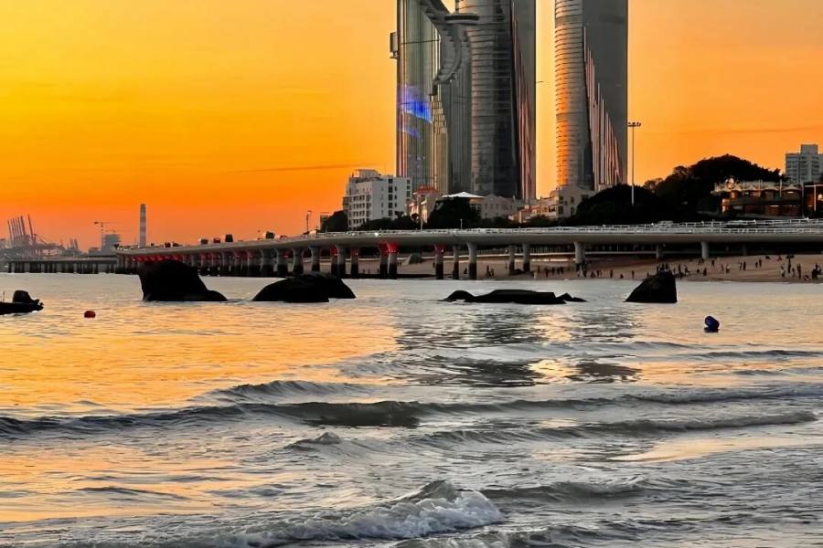 日落时从浅水处望向世茂海峡大厦双塔,天空橙红,前景是涌动的海浪与几块黑色礁石,横过画面的是演武大桥,桥后沙滩上有人影