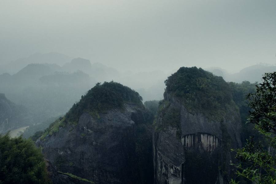 云雾中的丹霞山峰，两座圆顶岩体并立在灰白雾气里，左侧谷底露出一段弯曲的水面和树丛，前景有竹叶